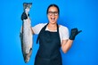 Young beautiful asian girl fishmonger selling fresh raw salmon pointing thumb up to the side smiling happy with open mouth
