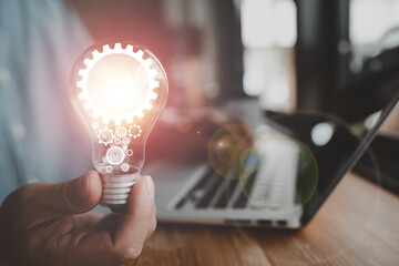 Poster - The hand of a man with a light bulb And there is a gear icon in it And he is using a notebook computer. The idea of ​​inspiration from online technology.innovation idea concept.