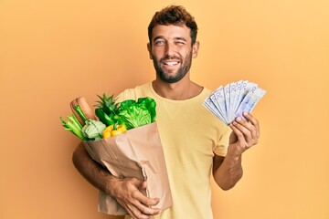 Wall Mural - Handsome man with beard holding groceries and 20 swedish krona banknotes winking looking at the camera with sexy expression, cheerful and happy face.