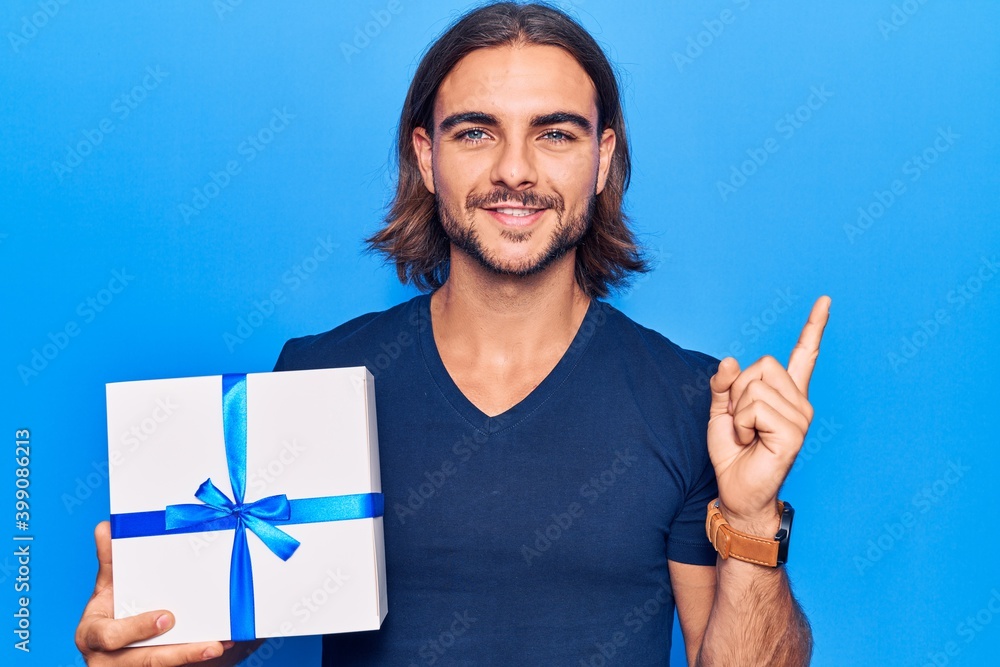 Young handsome man holding gift smiling happy pointing with hand and finger to the side