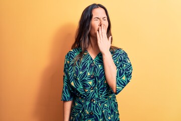 Wall Mural - Young beautiful brunette woman wearing casual floral dress standing over yellow background bored yawning tired covering mouth with hand. Restless and sleepiness.