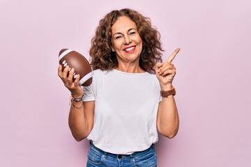 Poster - Middle age beautiful sporty woman playing rugby holding football ball over pink background smiling happy pointing with hand and finger to the side