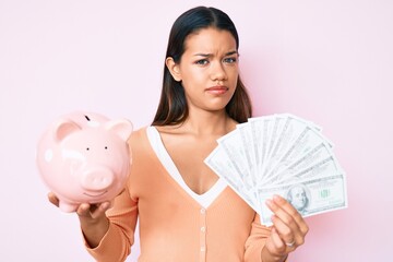 Sticker - Young beautiful latin girl holding piggy bank and dollars banknotes clueless and confused expression. doubt concept.