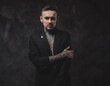 © Fxquadro - Invidivual and stylish guy with tattooed body and modern hairstyle in black suit poses in dark background looking at camera with serious face.