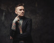 © Fxquadro - Dressed in black luxurious jacket serious and tattooed hipster person with modern haircut and short beard poses in dark background.