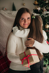 Young woman holding a present and hugging her daughter in front of the New Year tree. Families on New Year concept.