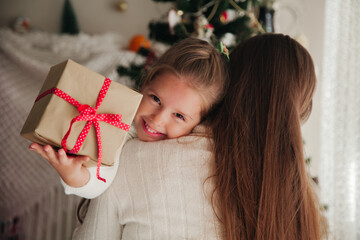 Wall Mural - Little girl holding a present and hugging her mum in front of the New Year tree. Families on New Year concept.
