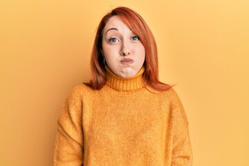 Sticker - Beautiful redhead woman wearing casual winter sweater over yellow background puffing cheeks with funny face. mouth inflated with air, crazy expression.