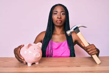 Wall Mural - African american woman with braids holding piggy bank and hammer relaxed with serious expression on face. simple and natural looking at the camera.