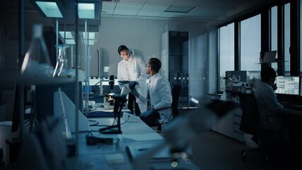 Modern Medical Research Laboratory: Two Scientists Working, Using Digital Tablet, Analyzing Test, Talking. Advanced Scientific Pharmaceutical Lab for Medicine, Biotechnology Development. Evening Time