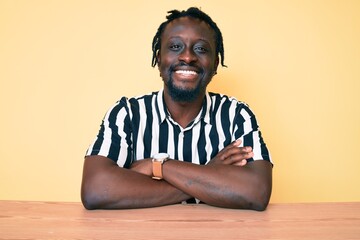 Sticker - Young african american man with braids wearing casual clothes sitting on the table happy face smiling with crossed arms looking at the camera. positive person.