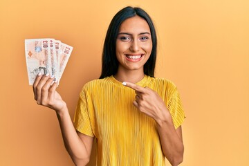 Poster - Young brunette woman holding 10 united kingdom pounds banknotes smiling happy pointing with hand and finger