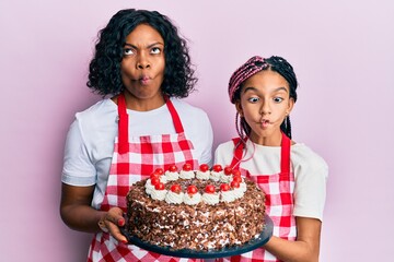 Wall Mural - Beautiful african american mother and daughter wearing baker apron holding homemade cake making fish face with mouth and squinting eyes, crazy and comical.