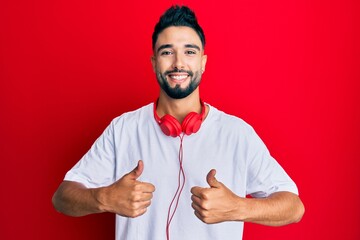 Wall Mural - Young man with beard listening to music using headphones success sign doing positive gesture with hand, thumbs up smiling and happy. cheerful expression and winner gesture.