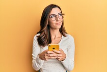 Young brunette woman using smartphone over yellow background smiling looking to the side and staring away thinking.