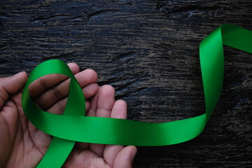 Top view of hands holding green color ribbon on dark background. Gall bladder and bile duct cancer, mental health, cerebral palsy awareness, organ donation, world kidney day and environmental care.