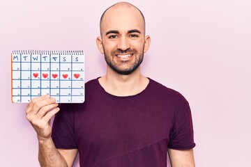 Young handsome bald man holding heart calendar looking positive and happy standing and smiling with a confident smile showing teeth