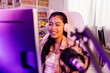 Excited and smiling gamer girl in cute headset with a mic playing an online video game and live streaming. Young Asian woman talking to players and audience on computer in neon led light room at home