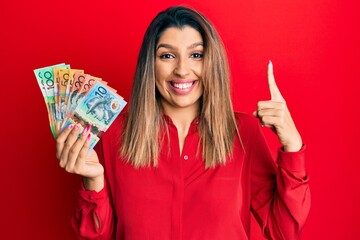 Poster - Beautiful brunette woman holding australian dollars smiling amazed and surprised and pointing up with fingers and raised arms.