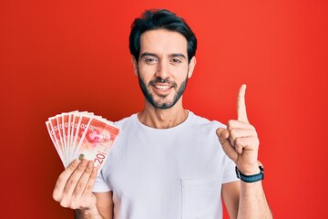 Sticker - Young hispanic man holding 20 israel shekels banknotes smiling with an idea or question pointing finger with happy face, number one