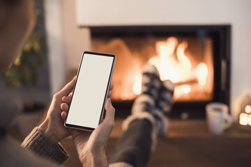 young woman using smart phone near fireplace at home, white blank empty screen display. winter lifes