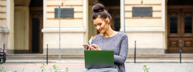 A young woman sits on the grass with a mobile phone and laptop computer against the background of a university