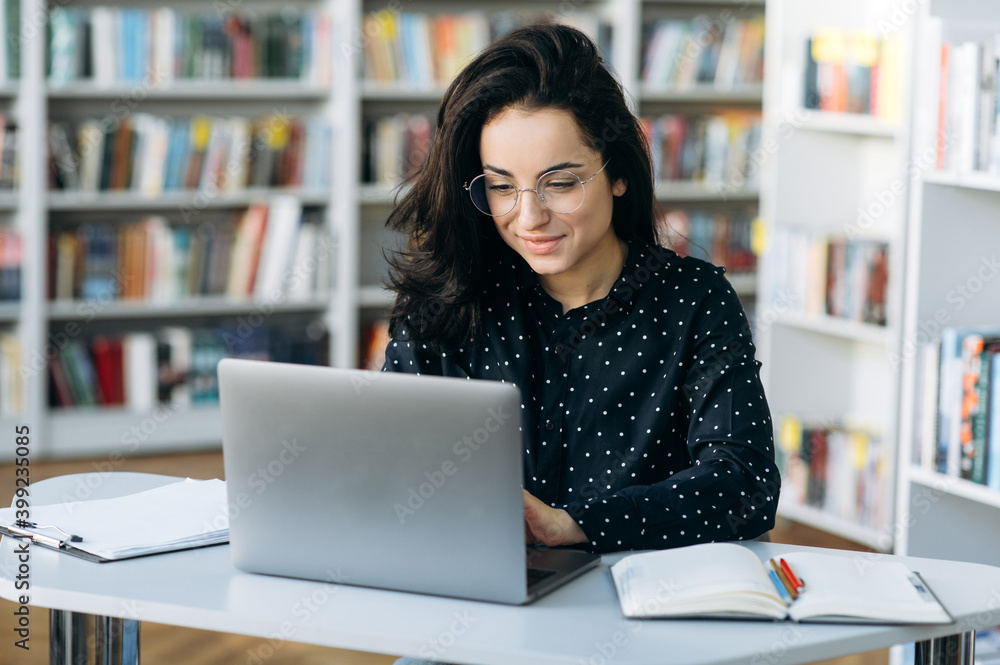 Busy caucasian young woman, in eyeglasses is sitting at the desk while remote work from home and chatting online using a laptop - obrazy, fototapety, plakaty 