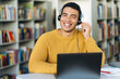 © Kateryna - Attractive hispanic young man, call center worker, student, or successful business man, in a headset is using a laptop while working online sitting at his desk and looking at the camera with smile