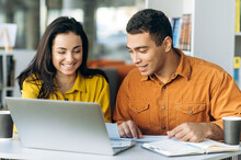 Two Colleagues Or Students, Caucasian Girl And Hispanic Guy, Are Using A Laptop While Sitting At Workplace Discussing About Job Or Education And Smiling