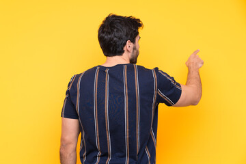 Wall Mural - Young handsome man with beard over isolated yellow background pointing back with the index finger