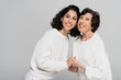 © LIGHTFIELD STUDIOS - Smiling hispanic woman holding hands of senior mother isolated on grey, two generations of women