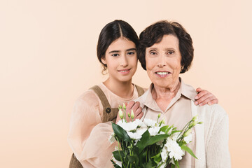 Smiling hispanic girl embracing granny with bouquet isolated on beige, two generations of women