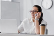 © LIGHTFIELD STUDIOS - Positive plus size hispanic businesswoman talking on smartphone at workplace with laptop on blurred background