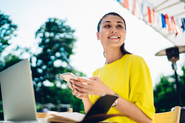 Wall Mural - Cheerful asian female in casual wear sitting in on cafe terrace working remotely using modern technology, smiling woman dialling number using mobile phone and laptop computer for remote job