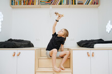 The boy plays with a wooden plane in the children's room, he sits on the ladder, raising his hand with the airplane above him