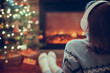 © flowertiare - Woman in headphones sitting and warming at winter evening near fireplace flame and  christmas tree.