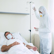© My Ocean studio - An adult male patient lies on a bed in a hospital ward while he is examined by a doctor in protective clothing and a mask.