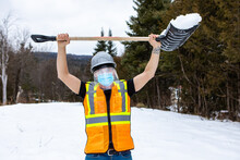 Medium Close Shot Of A Man Wearing An Orange Fluo Visibility Jacket, Covid Face Mask And Plastic Visor, Lifting A Shovel Of Snow Over His Head
