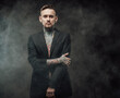 © Fxquadro - Invidivual and stylish guy with tattooed body and modern hairstyle in black suit poses in dark background looking at camera with serious face.