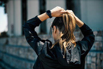 Rear view of young attractive, fit caucasian female runner standing in the urban exterior and tying ponytail.