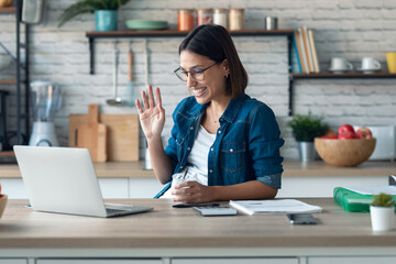 Wall Mural - Smiling young woman waving through the laptop web camera while eating yogurt in the kitchen at home.