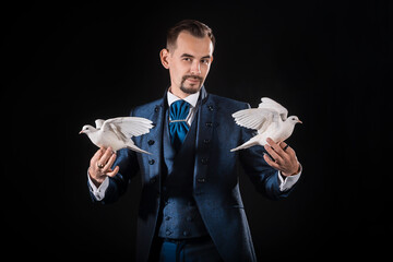 A magician in a blue tailcoat holds two white doves in his hands on a black background