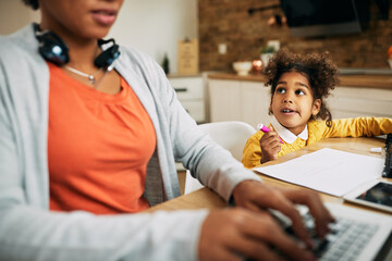 Wall Mural - Cute black girl drawing on the paper while her mother is working on laptop at home.