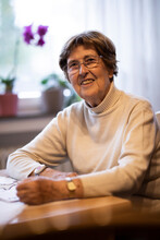 Smiling wrinkled woman with emergency button on wrist sitting by table at home