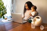 Fototapeta Tęcza - A beautiful young mixed race African American mother holds her daughter while taking notes at her dining table serving as a temporary remote work from home station with breast pump in foreground.