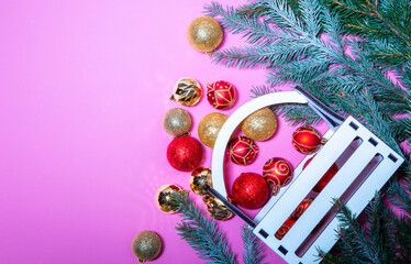 Christmas red and gold balls are scattered near a wooden box and fir trees on a pink background. View from above. space for text..