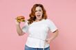 © ViDi Studio - Irritated young redhead plus size body positive woman 20s in white casual t-shirt hold american classic fast food burger measuring waist with measure tape isolated on pink background studio portrait.
