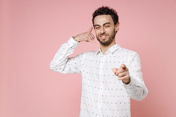 Wall Mural - Smiling young bearded man 20s wearing basic casual white shirt doing phone gesture like says call me back pointing index finger on camera isolated on pastel pink color wall background studio portrait.