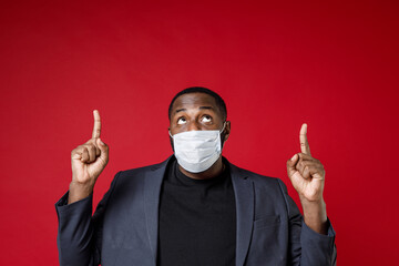 Young african american business man 20s in classic jacket suit sterile face mask to safe from coronavirus virus covid-19 pointing index fingers up isolated on red color background studio portrait.