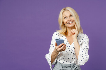 Pensive elderly gray-haired blonde woman lady 40s 50s years old in white dotted blouse using mobile phone typing sms message put hand prop up on chin isolated on violet background studio portrait.